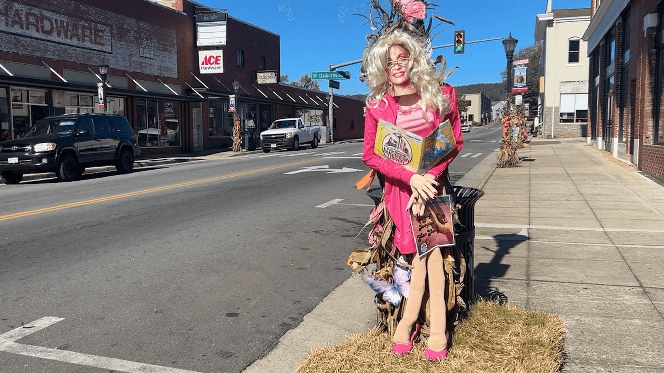 A Dolly Parton scarecrow with a blonde wig and pink outfit, holding a book, on a Rocky Mount sidewalk